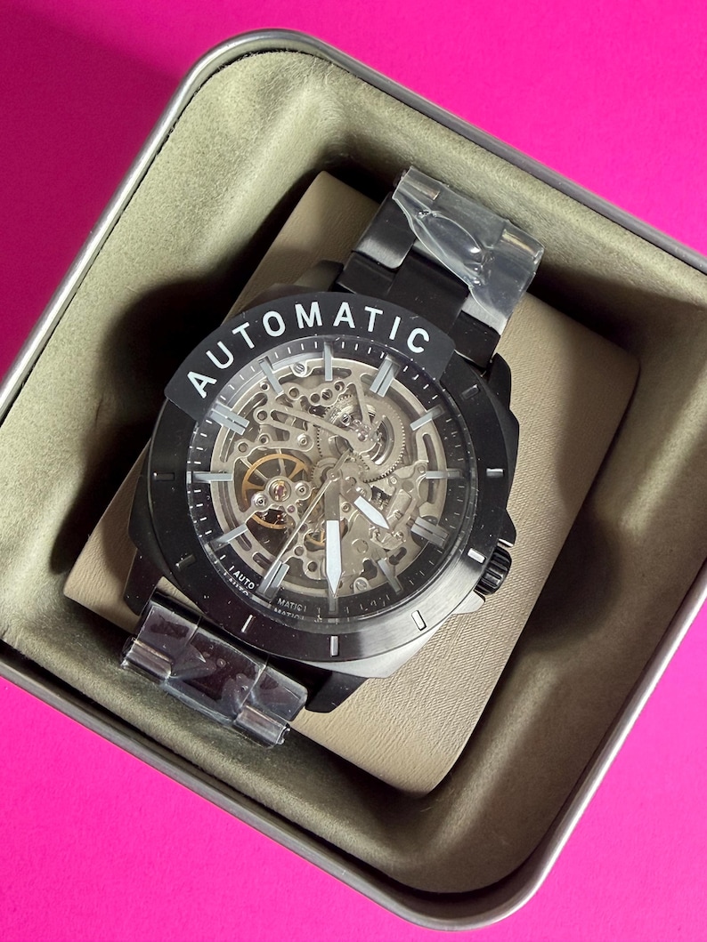 May include: Black automatic watch with a skeleton dial, visible gears, and white hands. The watch is in a metal case with the word "AUTOMATIC" on the bezel. The watch is new, with protective plastic on the band.