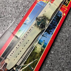Op de afbeelding: Een wit plastic horloge met een helder gezicht en een zwart en zilver ontwerp. Het horloge bevindt zich in een transparante plastic display met een rode en witte achtergrond.