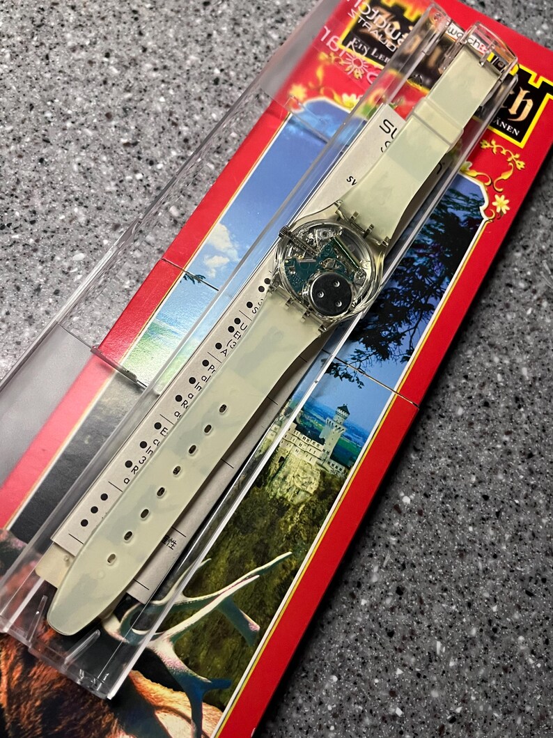 Op de afbeelding: Een wit plastic horloge met een helder gezicht en een zwart en zilver ontwerp. Het horloge bevindt zich in een transparante plastic display met een rode en witte achtergrond.