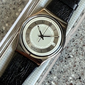 K&ouml;nnte beinhalten: Eine silberfarbene Swatch-Uhr mit schwarzem Lederarmband. Das Zifferblatt der Uhr ist wei&szlig; mit schwarzen r&ouml;mischen Ziffern und Zeigern. Die Uhr befindet sich in der Originalverpackung.