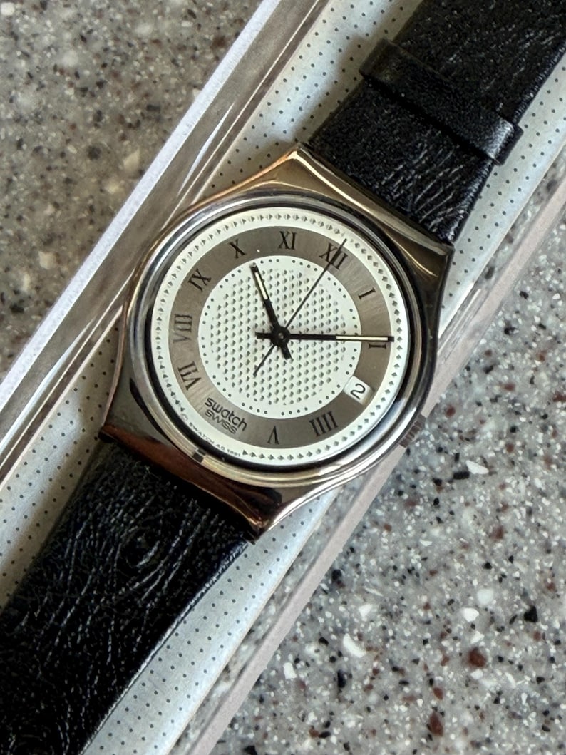 K&ouml;nnte beinhalten: Eine silberfarbene Swatch-Uhr mit schwarzem Lederarmband. Das Zifferblatt der Uhr ist wei&szlig; mit schwarzen r&ouml;mischen Ziffern und Zeigern. Die Uhr befindet sich in der Originalverpackung.