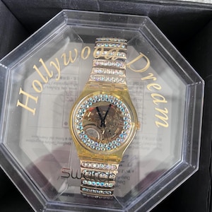 May include: A gold-tone watch with a clear face and a crystal-encrusted band. The watch is in a clear plastic box with the words "Hollywood Dream" printed on the side. The watch is a Swatch brand.
