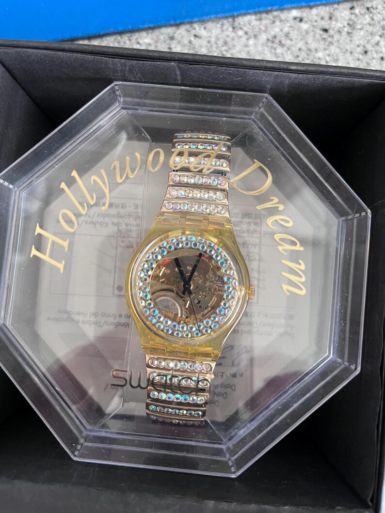 May include: A gold-tone watch with a clear face and a crystal-encrusted band. The watch is in a clear plastic box with the words "Hollywood Dream" printed on the side. The watch is a Swatch brand.