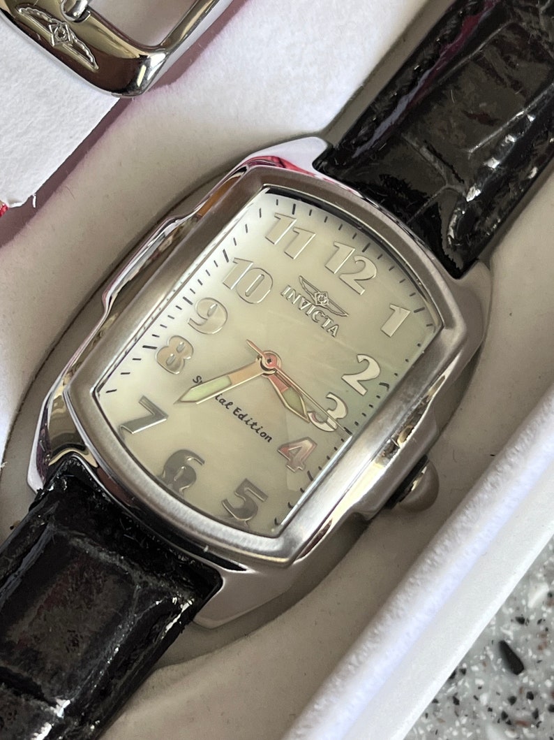 Puede incluir: Un reloj Invicta plateado con una esfera blanca y una correa de cuero negra. La esfera del reloj tiene marcadores de hora romanos negros y una aguja de segundos plateada. La esfera del reloj tambi&eacute;n tiene el texto "Invicta" y "Special Edition".