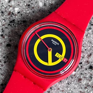 Può includere: Un orologio Swatch rosso con quadrante nero e giallo. Il quadrante mostra il giorno della settimana e la data. L'orologio ha un cinturino rosso e un quadrante rotondo. L'orologio è al quarzo.