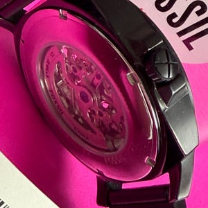 May include: Close-up of a black Fossil watch with a skeleton dial, showcasing the inner workings. The watch has a black metal band and a pink background. The watch face is visible, with the brand name "Fossil" partially visible.