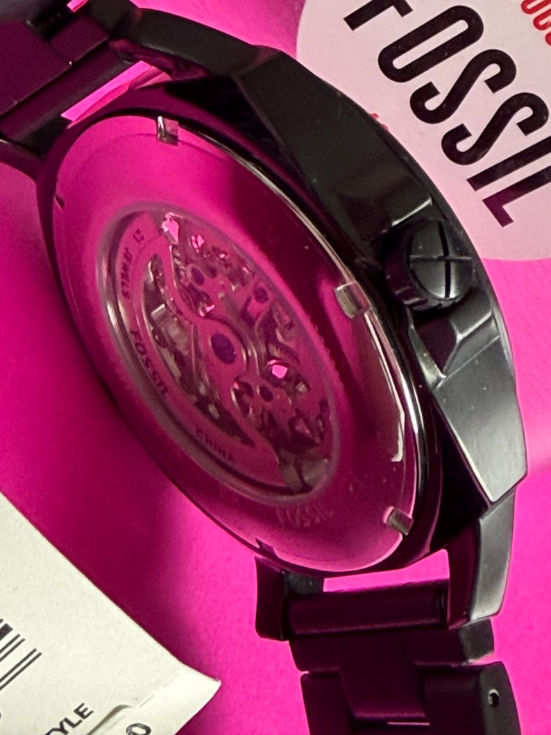 May include: Close-up of a black Fossil watch with a skeleton dial, showcasing the inner workings. The watch has a black metal band and a pink background. The watch face is visible, with the brand name "Fossil" partially visible.
