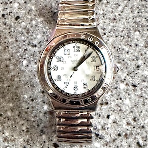 May include: A silver-toned Swatch watch with a white face and black numerals. The watch features a round dial with a second time indicator and a silver band. The watch hands point to 1:07.