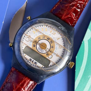 May include: A Swatch watch with a white face, black bezel, and a red leather band. The watch face displays the time with gold accents and a digital display. The band has a textured, crocodile-like pattern.