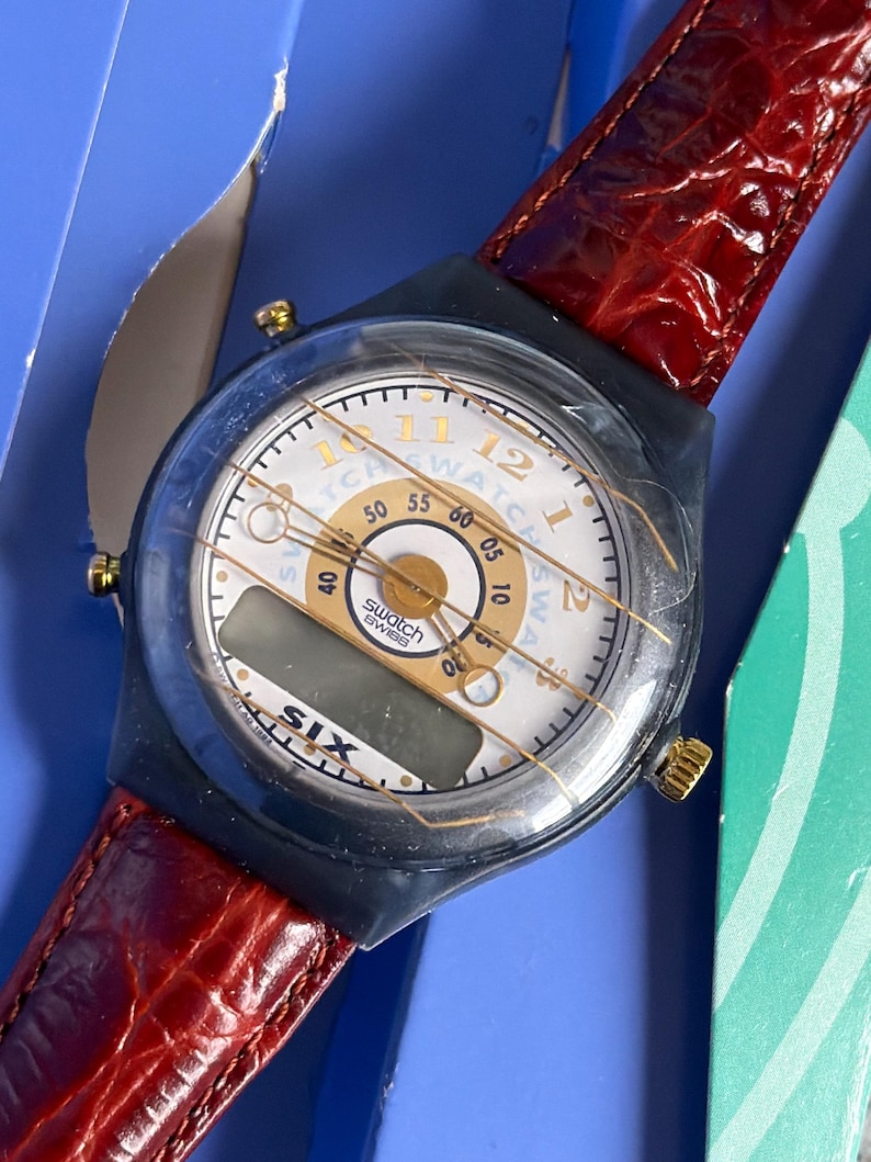 May include: A Swatch watch with a white face, black bezel, and a red leather band. The watch face displays the time with gold accents and a digital display. The band has a textured, crocodile-like pattern.