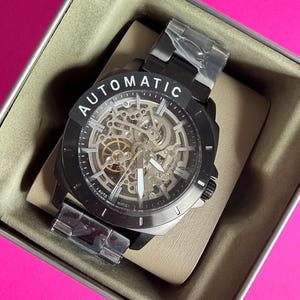 May include: A black automatic watch with a skeleton dial, displayed in a tan box. The watch features a black bezel and band, with the word "AUTOMATIC" printed on the face. The watch is new, with protective plastic on the band.
