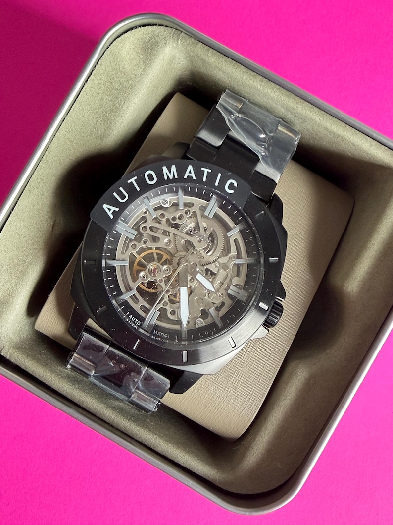 May include: A black automatic watch with a skeleton dial, displayed in a tan box. The watch features a black bezel and band, with the word "AUTOMATIC" printed on the face. The watch is new, with protective plastic on the band.