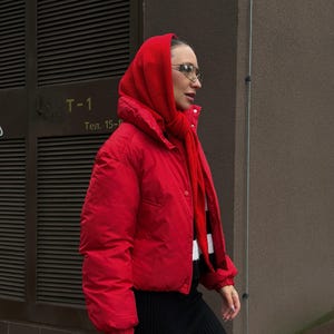 以下が含まれることがあります： 赤いフード付きスカーフ、ジャケットを着て、赤いハンドバッグを持っている人。服装には黒いスカートと白と黒のストライプのセーターが含まれています。その人はまた、メガネをかけています。
