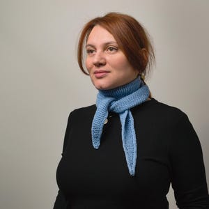 Op de afbeelding: Een vrouw met rood haar draagt een zwart shirt met lange mouwen en een lichtblauwe gebreide sjaal om haar nek.