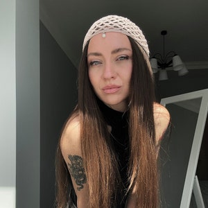 May include: A woman with long brown hair wearing a beige crocheted hat with a pearl accent. She is wearing a black mock neck shirt and has a dragon tattoo on her upper arm.