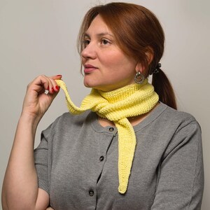 May include: A woman wearing a gray shirt with a yellow crocheted scarf around her neck. She is holding the end of the scarf in her right hand.