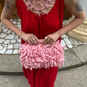 May include: A bright pink, handmade clutch with a textured, looped design. The clutch is held against a red dress, and a coral-like necklace is worn around the neck. Several rings adorn the fingers.