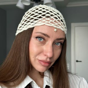 May include: A white crochet hat with a pearl accent. The hat is worn by a person with long brown hair.