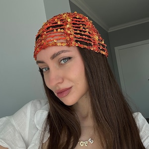 Pode incluir: Uma mulher usando um chapéu vermelho e dourado com lantejoulas. O chapéu tem um design de malha e é usado sobre seu cabelo castanho longo. Ela está usando uma camisa branca e um colar de ouro com a palavra "LOVE" nele.