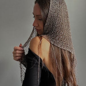 May include: A shimmering, open-weave head covering in a neutral tone, draped over the head and shoulders. The delicate fabric features a net-like pattern with dangling strands. The garment is worn over a black long-sleeved top.