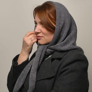 May include: A woman wearing a gray hooded scarf and a dark gray coat. The scarf is made of a soft, knitted fabric and is pulled up over her head.