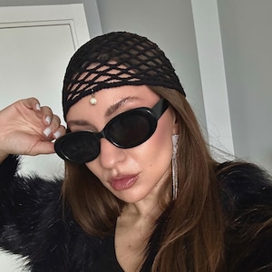 May include: A woman wearing a black fishnet hair covering, black oval sunglasses, and long dangling earrings. She is wearing a black fur jacket.