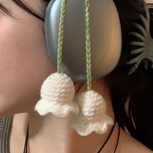 May include: A pair of blue headphones with white crocheted bell charms attached to the headband. The charms are hanging down on either side of the headphones.
