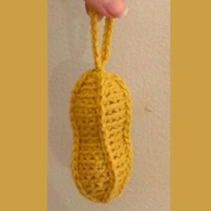 Puede incluir: Un adorno de cacahuete tejido a crochet en color amarillo. El adorno tiene un lazo en la parte superior para colgarlo.