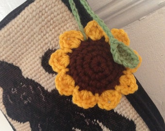 Puede incluir: Una bolsa de crochet beige con una silueta negra de un gato y un girasol de crochet amarillo con un centro marrón.