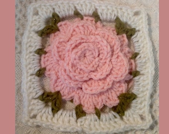 May include: A crocheted square with a pink flower in the center. The flower is surrounded by a white border with green accents.
