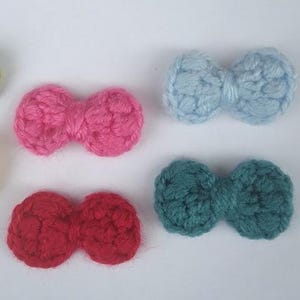 May include: A collection of six handmade crochet bow accents in various colors. The bows are in shades of green, yellow, pink, red, blue, and teal. These small, decorative bows are likely used for crafting or embellishment purposes.