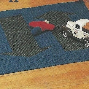 Op de afbeelding: Een blauw en groen tapijt met een groot "L"-ontwerp. Een speelgoedvrachtwagen met een witte cabine en bruine lading ligt op het tapijt. Er ligt ook een rood en blauw speeltje op het tapijt. Het tapijt ligt op een houten vloer.