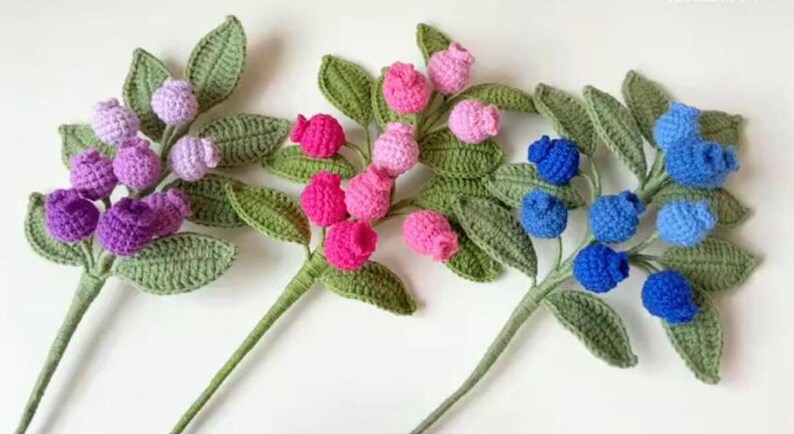 May include: Three crocheted flower stems with green leaves and purple, pink, and blue berries.