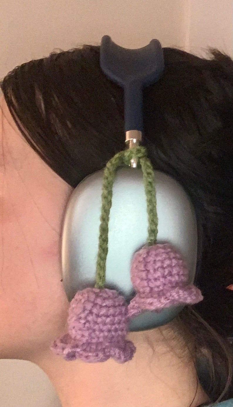 May include: A pair of purple crocheted flower ear warmers with green yarn strings attached to a blue headband.