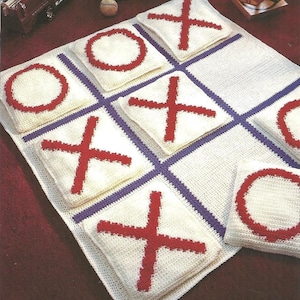 May include: A crocheted tic-tac-toe game board featuring white squares, purple lines, and red crosses and noughts. The game is on a red surface, with toy cars and other toys in the background.