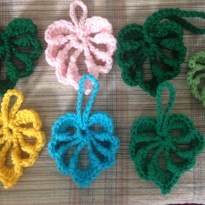 May include: A selection of handmade crochet leaf decorations in shades of green, pink, yellow, and blue. Each leaf features a hanging loop and detailed openwork, ideal for seasonal displays. Measurements are in centimetres.