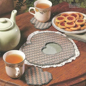 Può includere: Una teiera in ceramica verde chiaro, due tazze bianche riempite di tè, un piatto di biscotti alla marmellata e sottobicchieri a forma di cuore con un motivo intrecciato grigio e rosa. I sottobicchieri presentano un bordo in pizzo bianco.