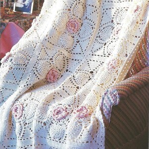 May include: Cream-colored, crocheted blanket with a floral pattern and pink rose accents. The blanket is draped over a chair, showcasing its intricate design and texture. A framed photo and candles are visible in the background.