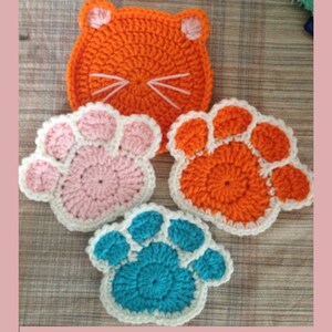 May include: A collection of handmade crochet items, including an orange cat face and three paw-shaped coasters. The coasters are in shades of pink, orange, and teal, each with a white border. The cat face has white whiskers.