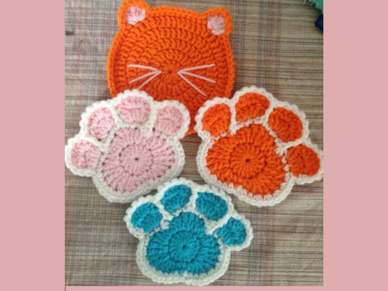 May include: A collection of handmade crochet items, including an orange cat face and three paw-shaped coasters. The coasters are in shades of pink, orange, and teal, each with a white border. The cat face has white whiskers.