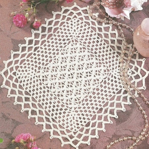 May include: A white, crocheted doily with a diamond-shaped design. The intricate lace pattern features a central motif surrounded by a scalloped edge. The doily is displayed on a mauve surface, with pink flowers and a beaded necklace in the background.
