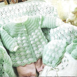 Puede incluir: Un conjunto de bebé de ganchillo verde menta, que incluye una rebeca, un gorro y patucos, con una manta a juego. El conjunto presenta un diseño de puntada de concha texturizada. La ropa de bebé se exhibe sobre una superficie blanca.