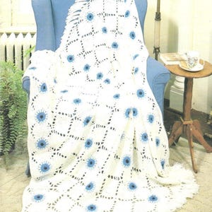 Könnte beinhalten: Eine weiße Häkeldecke mit blauem Blumenmuster, drapiert über einem blauen Sessel. Die Decke hat einen Fransenrand und ist aus weichem, strukturiertem Garn gefertigt. Das Blumenmuster ist gleichmäßig über die Decke verteilt.