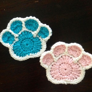 May include: Two handmade crochet paw print coasters. One is turquoise with a white border, and the other is pink with a white border. The coasters are made of yarn and are designed to protect surfaces from drink condensation.