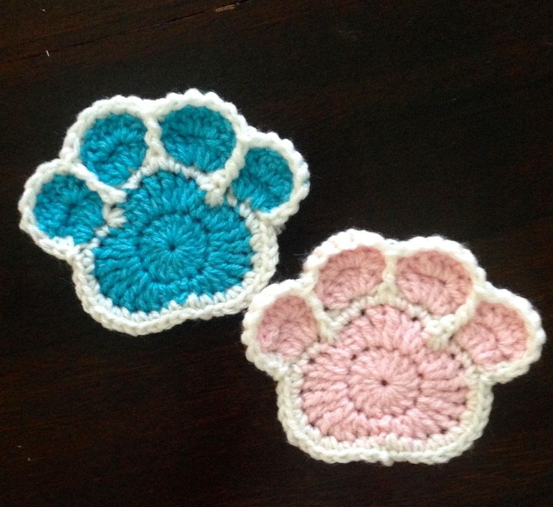 May include: Two handmade crochet paw print coasters. One is turquoise with a white border, and the other is pink with a white border. The coasters are made of yarn and are designed to protect surfaces from drink condensation.