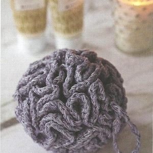 Può includere: Primo piano di una spugna da bagno all'uncinetto color lavanda con un anello per appenderla. La spugna ha un design strutturato e ad anello. Due tubetti di lozione e una candela accesa sullo sfondo.