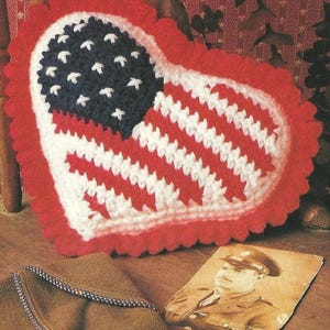 Puede incluir: Un cojín en forma de corazón, tejido a ganchillo, con el diseño de la bandera estadounidense. El cojín tiene un borde rojo festoneado, un campo azul con estrellas blancas y rayas rojas y blancas. También se ve una fotografía en tonos sepia y un paño marrón doblado.