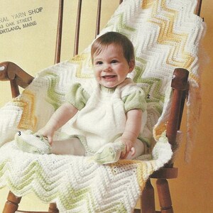 May include: A baby sits in a wooden rocking chair, smiling. The baby is wearing a white dress with green sleeves and matching shoes. A handmade, chevron-patterned blanket in white, yellow, and green covers the chair.