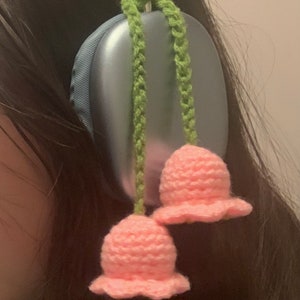 May include: A pair of blue headphones with pink crocheted flower charms attached to the headband. The charms are hanging down from green crocheted cords.
