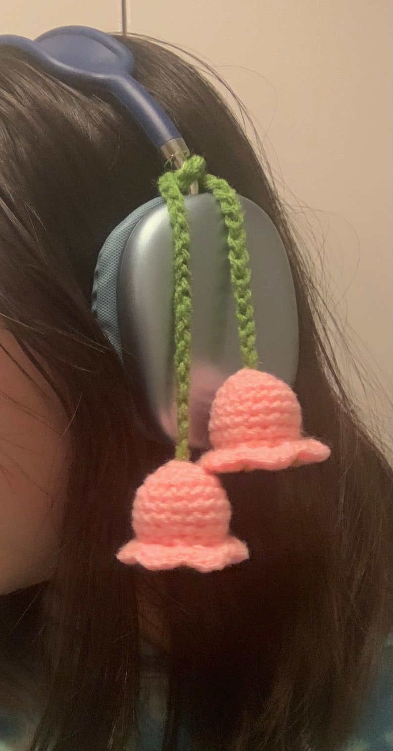 May include: A pair of blue headphones with pink crocheted flower charms attached to the headband. The charms are hanging down from green crocheted cords.